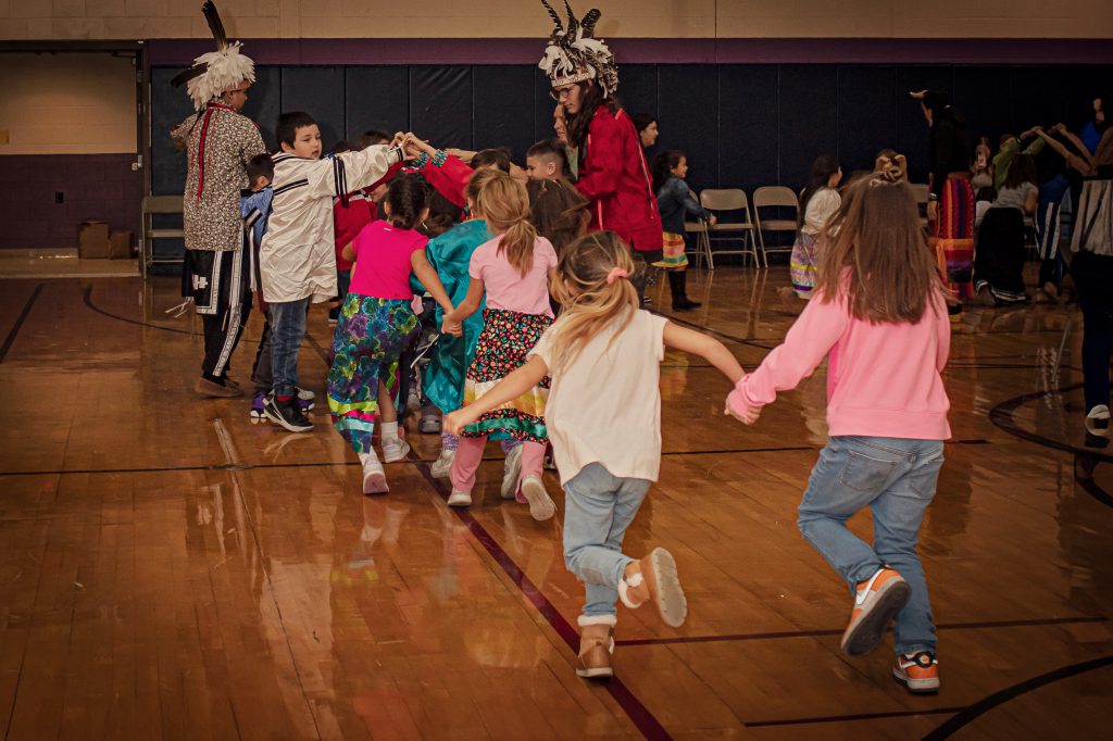 Several young students running in a gym with some older students wearing native attire