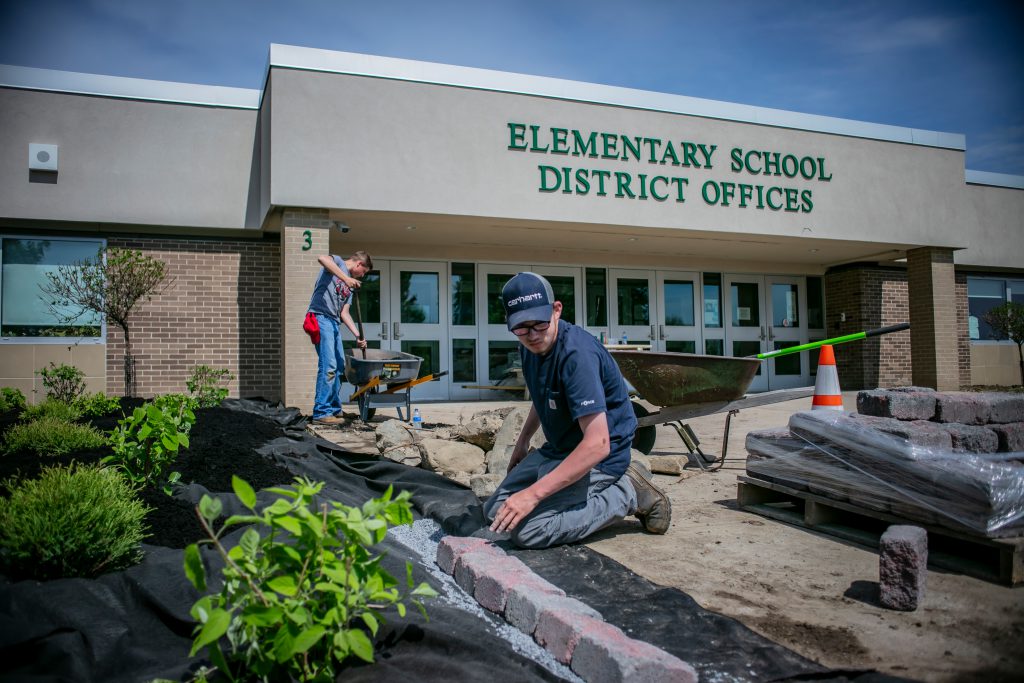 A student kneeling before a garden laying brick with another student in the background shoveling in a wheel barrow