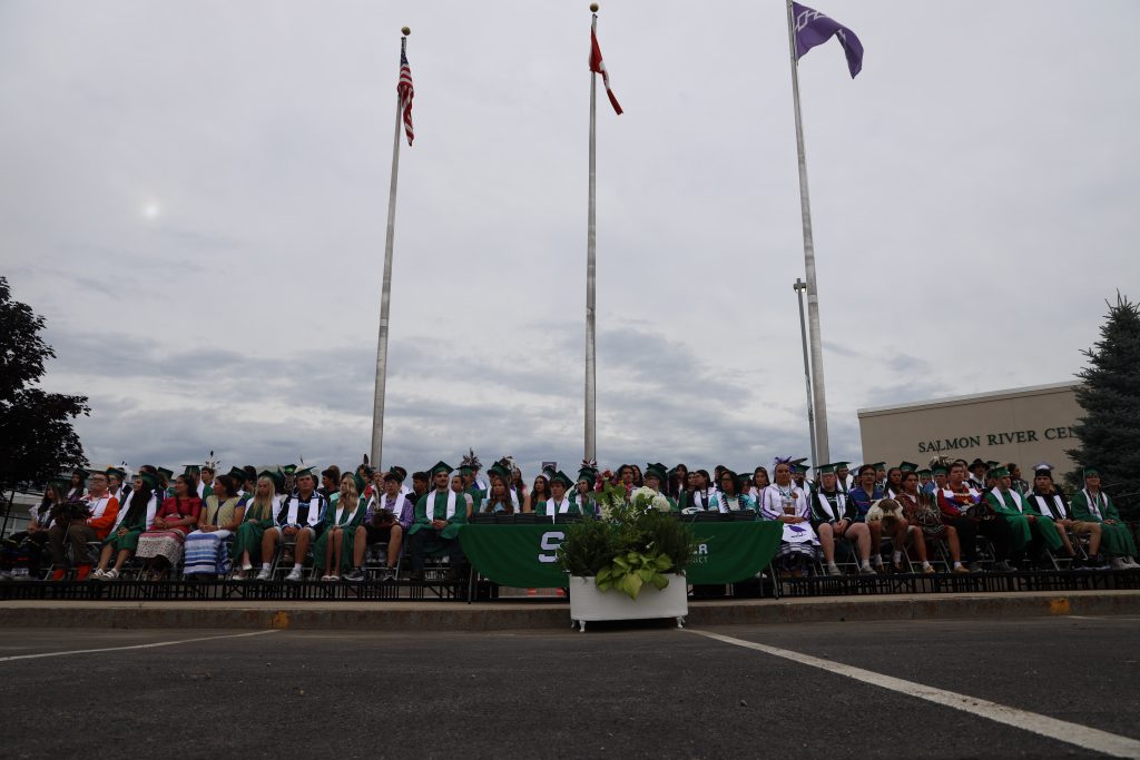 Dozens of students sitting in chairs wearing graduation attire with three flags flying high above them