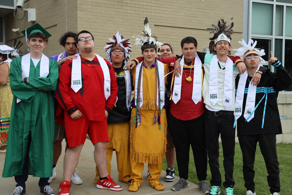 A group of graduates, each wearing white sashes stand together outside the school