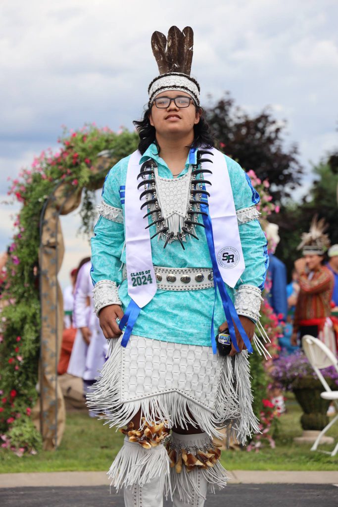 A graduate in native dress and claw necklace walks toward the camera