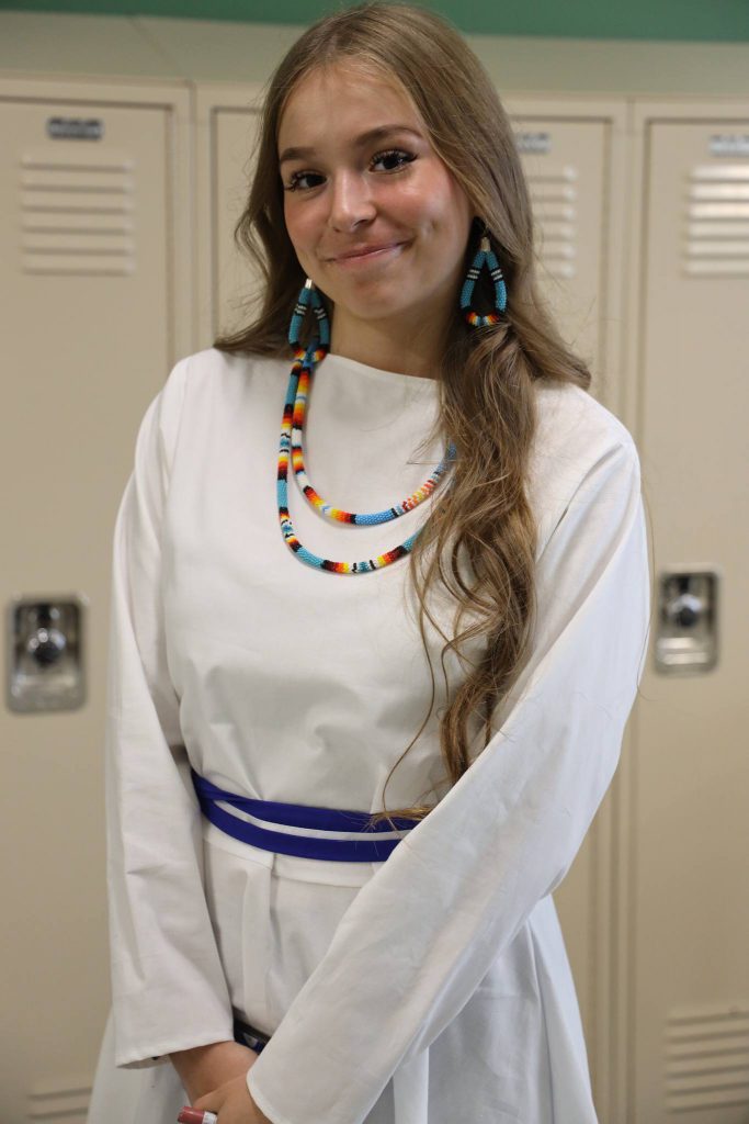 One student in white dress and native jewelry smiles in front of school lockers