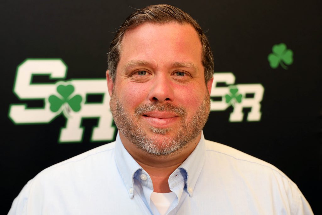 Man smiling in front of a back drop with the letters SR on it