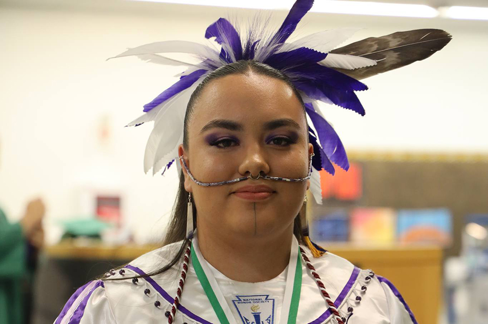 A high school student wears a feather headdress and facial jewelry