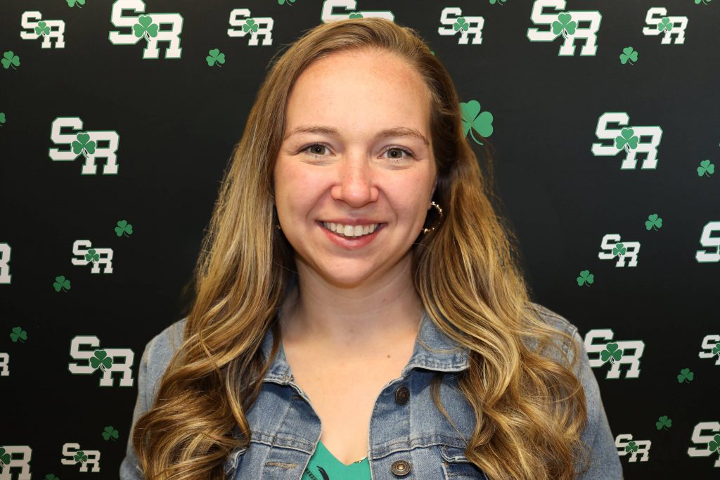 A woman smiling for a photo with a black background with green highlights with the letters 'SR'