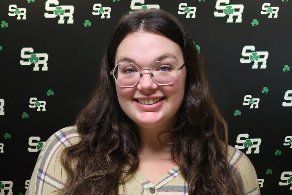 A woman smiling for a photo with a black background with green highlights with the letters 'SR'