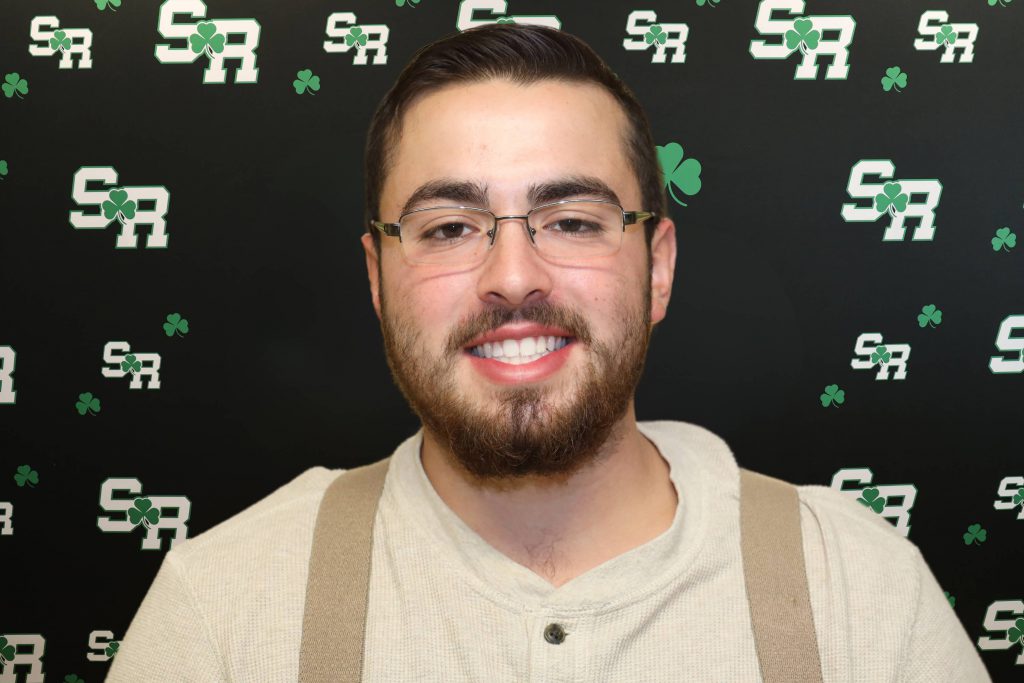 A man smiling for a photo with a black background with green highlights with the letters 'SR'