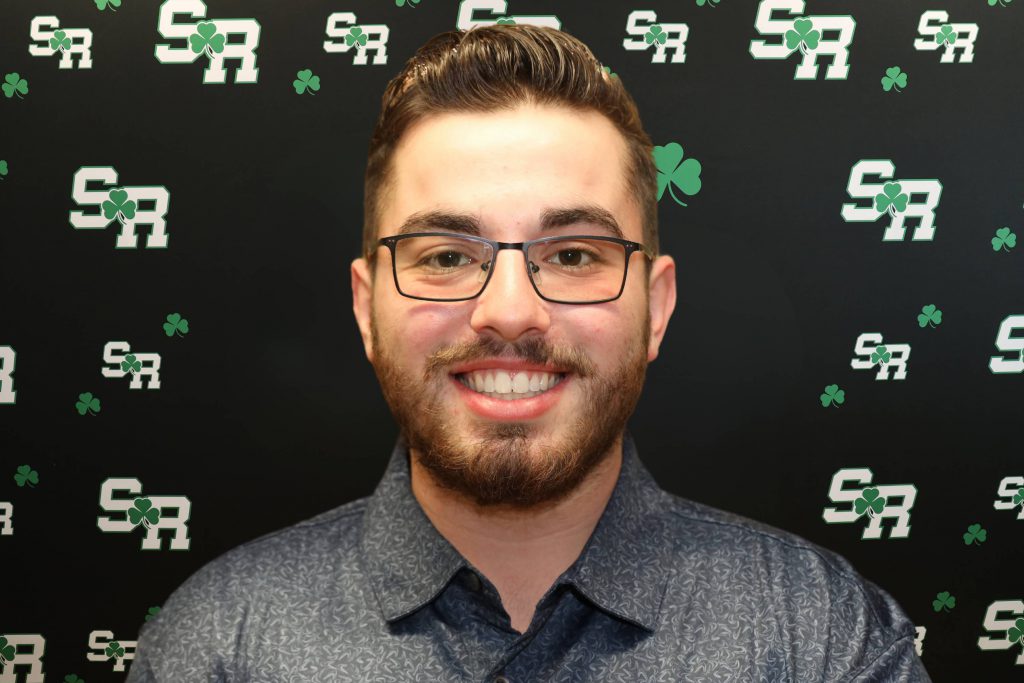 A man smiling for a photo with a black background with green highlights with the letters 'SR'