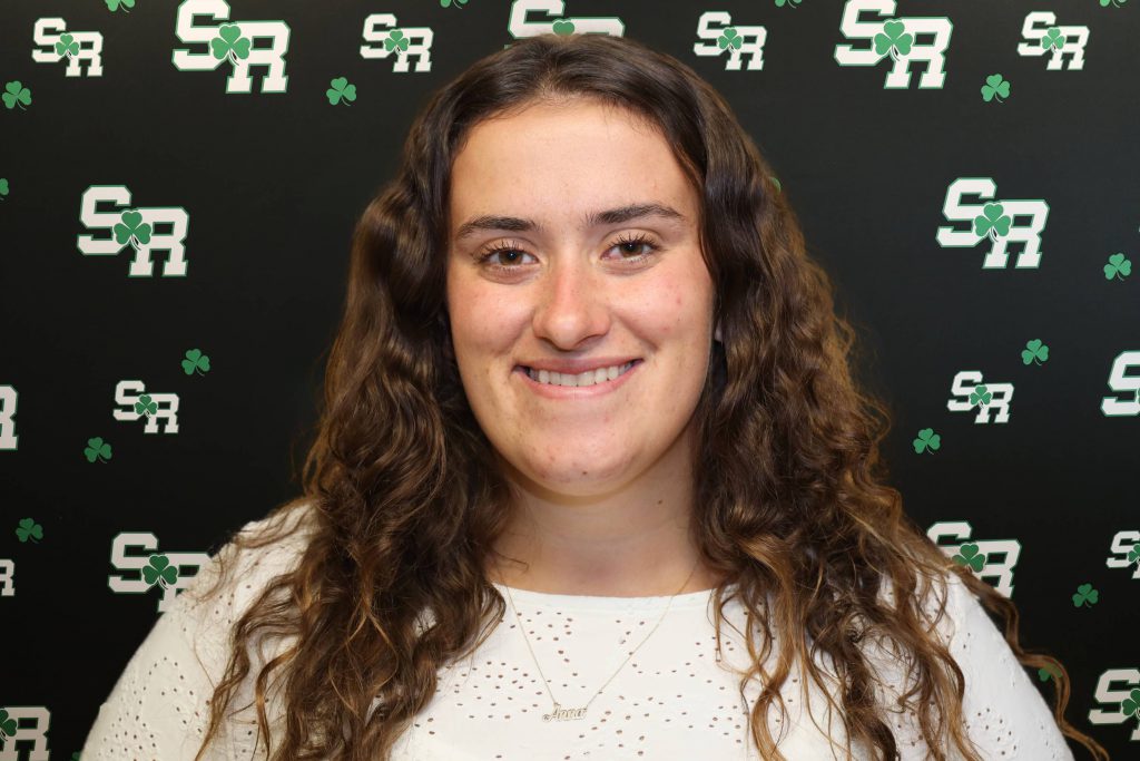 A woman smiling for a photo with a black background with green highlights with the letters 'SR'