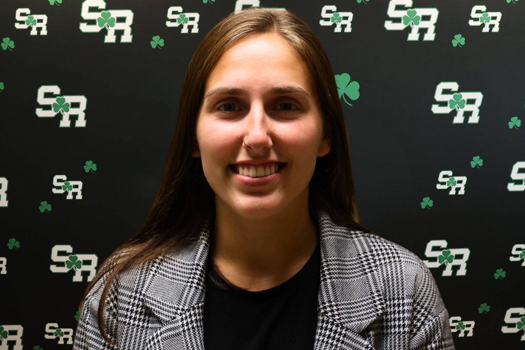 A woman smiling for a photo with a black background with green highlights with the letters 'SR'