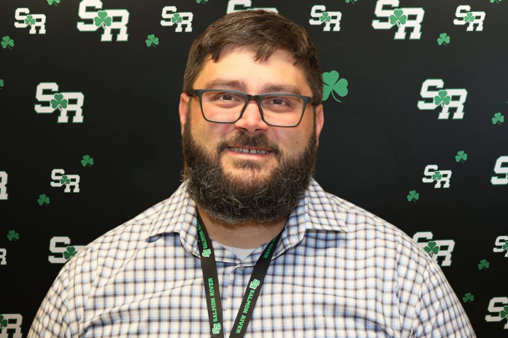 A man smiling for a photo with a black background with green highlights with the letters 'SR'