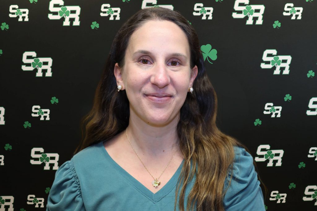 A woman smiling for a photo with a black background with green highlights with the letters 'SR'