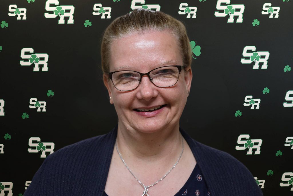 A woman smiling for a photo with a black background with green highlights with the letters 'SR'