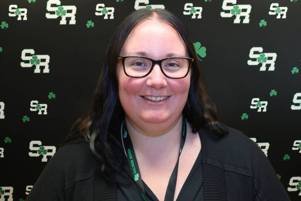 A woman smiling for a photo with a black background with green highlights with the letters 'SR'