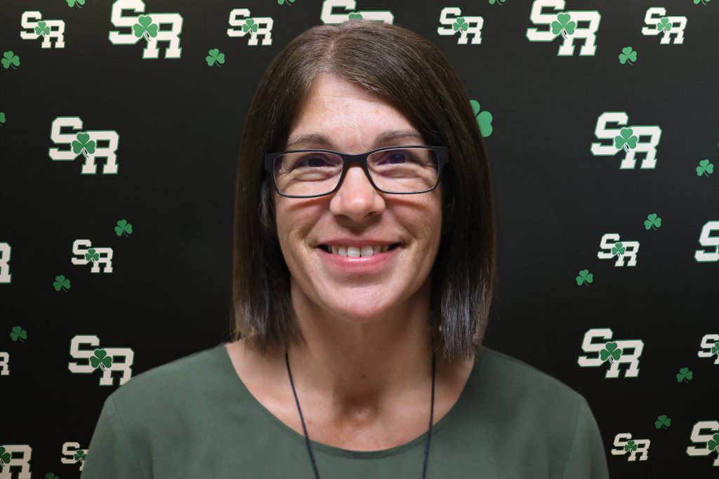 A woman smiling for a photo with a black background with green highlights with the letters 'SR'