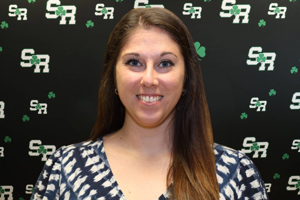 A woman smiling for a photo with a black background with green highlights with the letters 'SR'