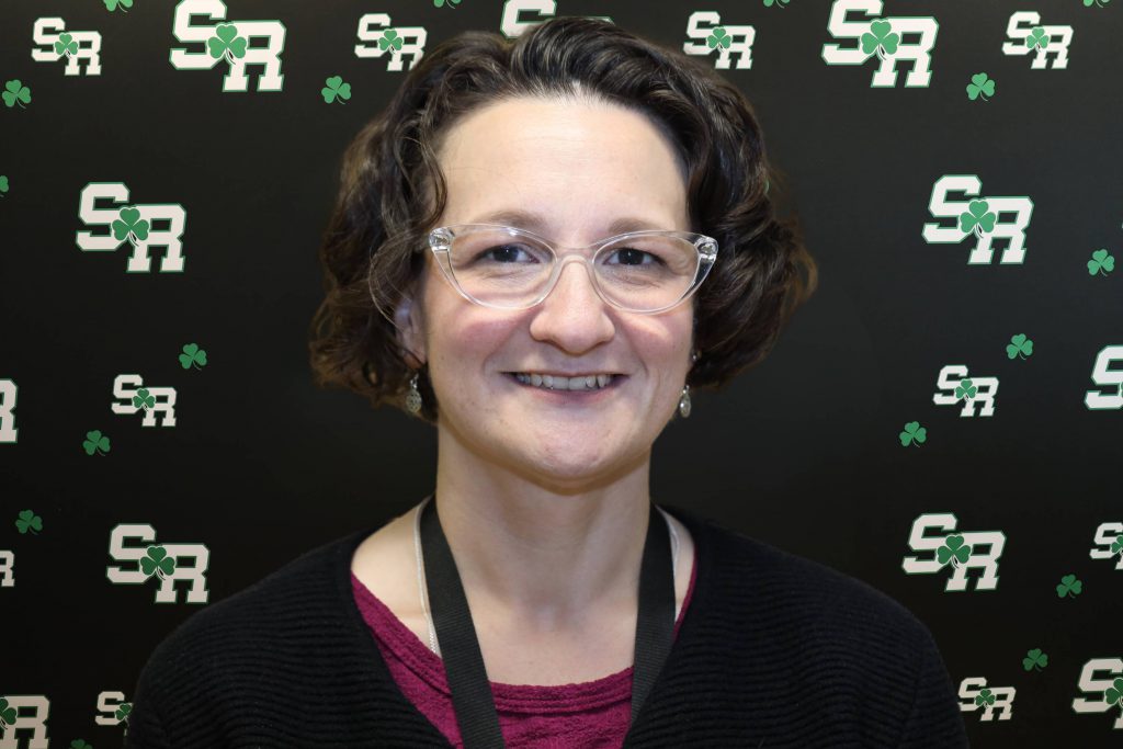 A woman smiling for a photo with a black background with green highlights with the letters 'SR'