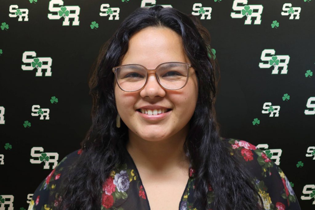 A woman smiling for a photo with a black background with green highlights with the letters 'SR'