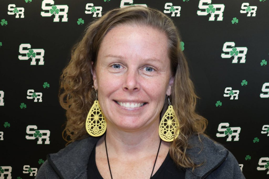 A woman smiling for a photo with a black background with green highlights with the letters 'SR'