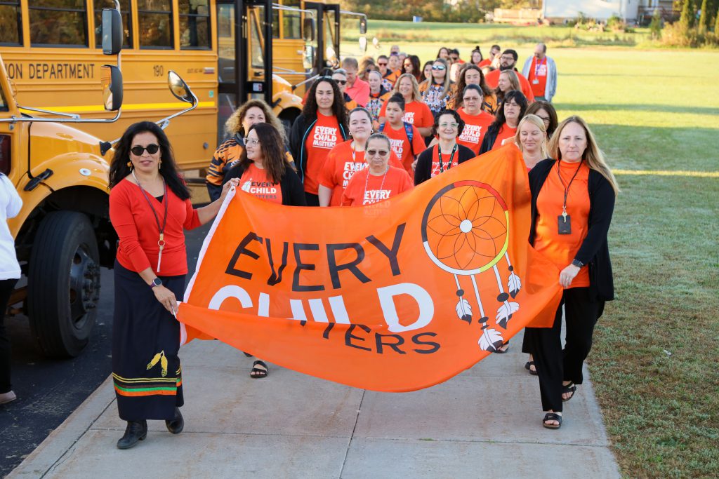 A group of adults wearing orange shirts walk along a sidewalk with two people in the front holding a banner that says 'Every Child Matters.'