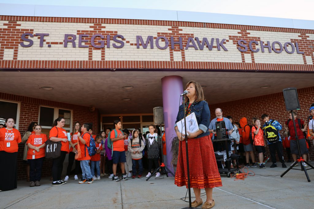 A woman standing at a microphone in front of a crowd of people in front of St. Regis Mohawk School