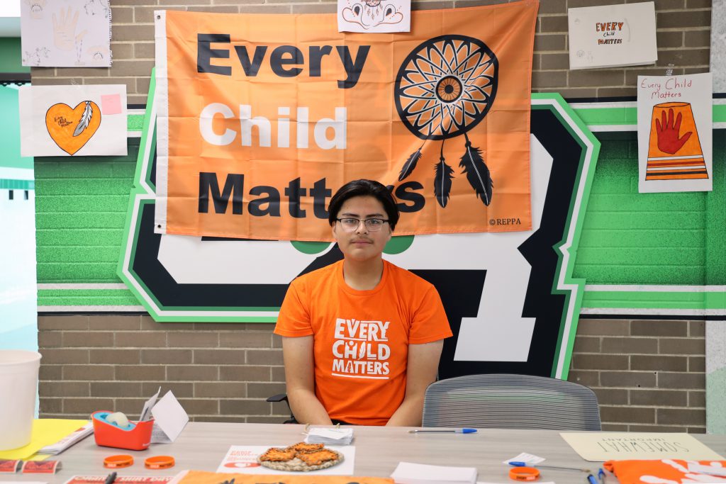 A student sits behind a table with a flag above him that says 'Every Child Matters'