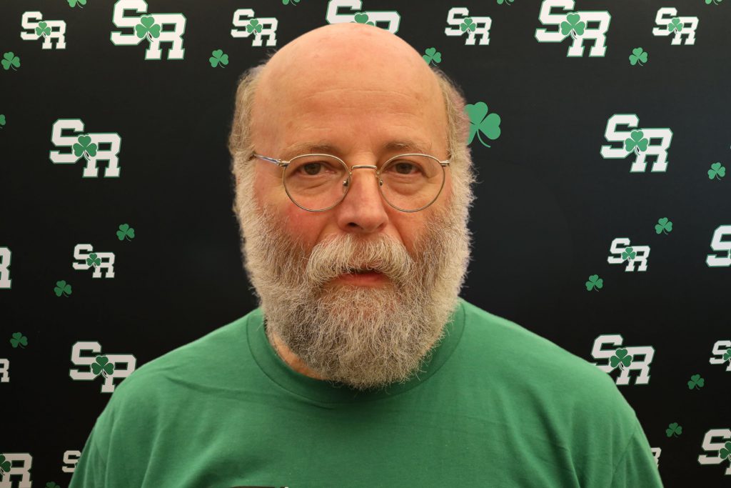 A man smiling for a photo with a black background with green highlights with the letters 'SR'