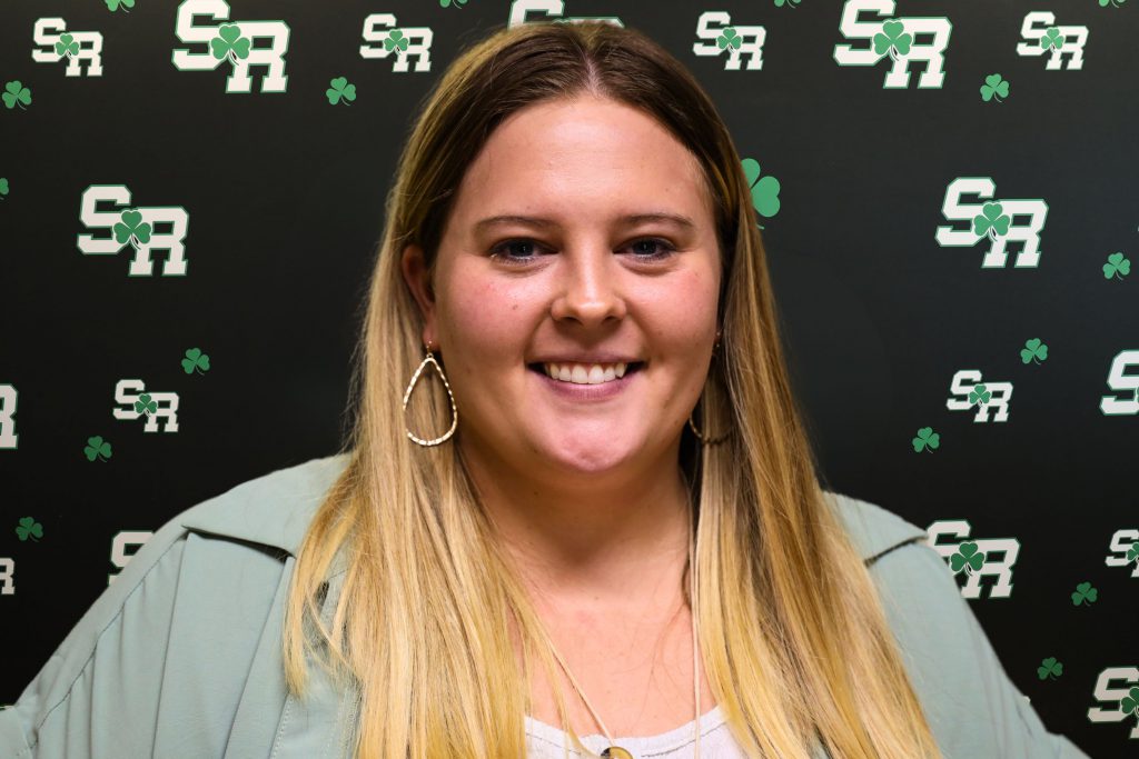 A woman smiling for a photo with a black background with green highlights with the letters 'SR'