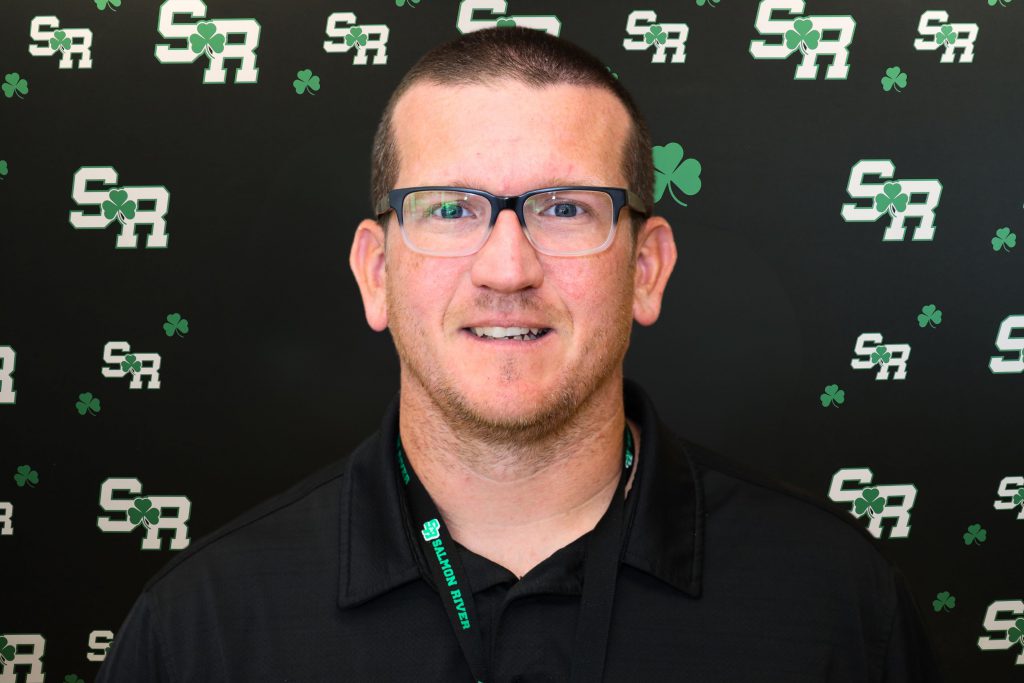 A man smiling for a photo with a black background with green highlights with the letters 'SR'