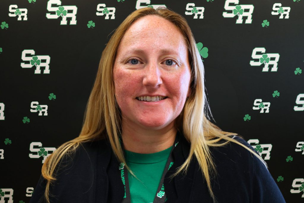 A woman smiling for a photo with a black background with green highlights with the letters 'SR'