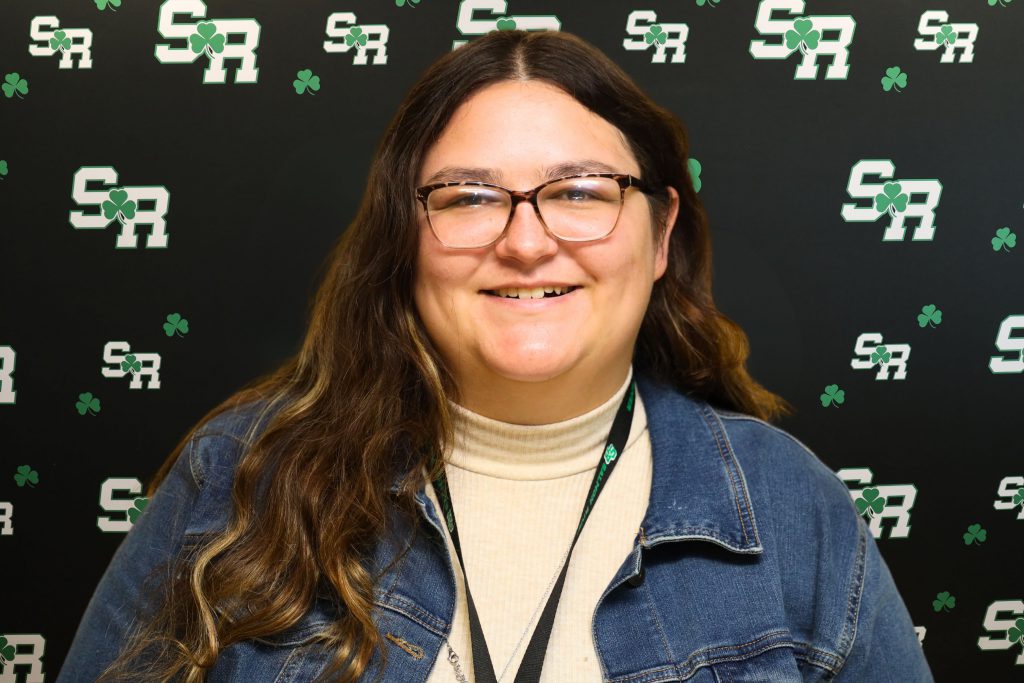 A woman smiling for a photo with a black background with green highlights with the letters 'SR'