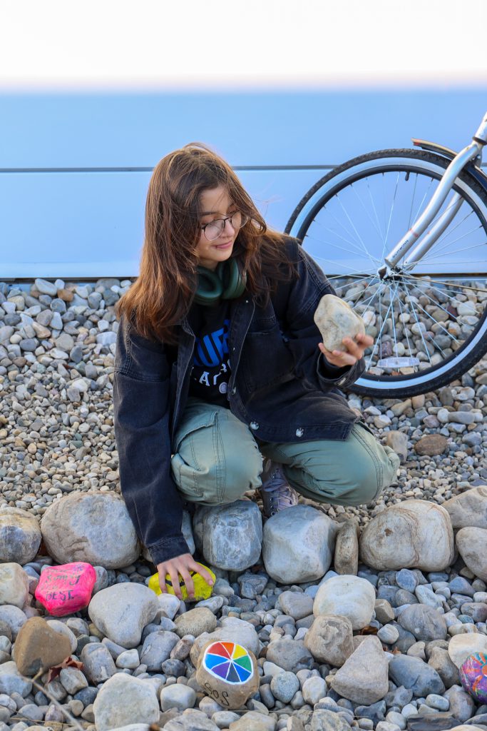 A student kneeling on a bed of rocks, holding up one painted rock