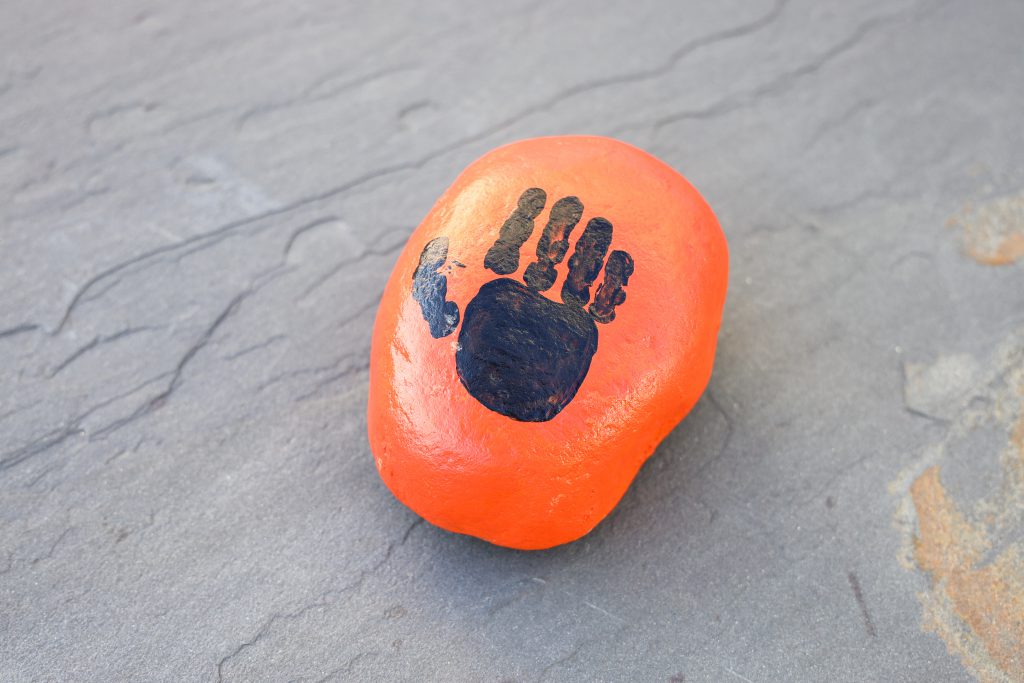 A closeup photo of a orange painted rock with the silhouette of a hand painted on it