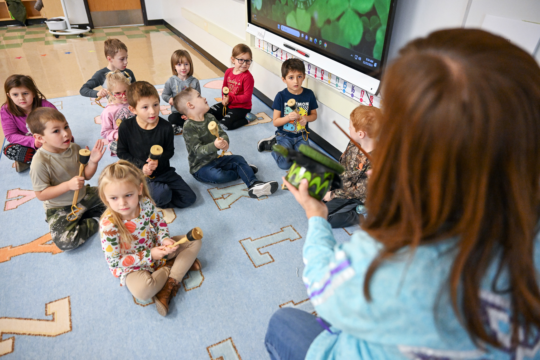 A teacher sits in front of a group of students who are sitting on the floor with instruments