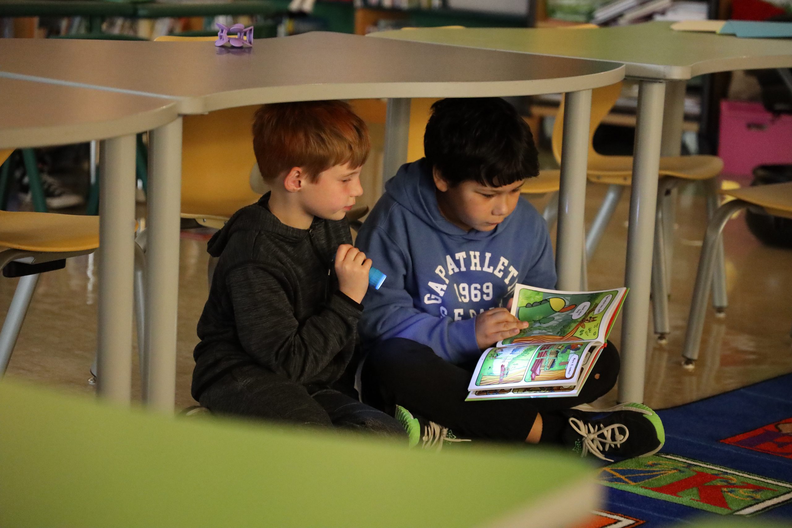 Two students sitting underneath a table reading a book together using a flashlight