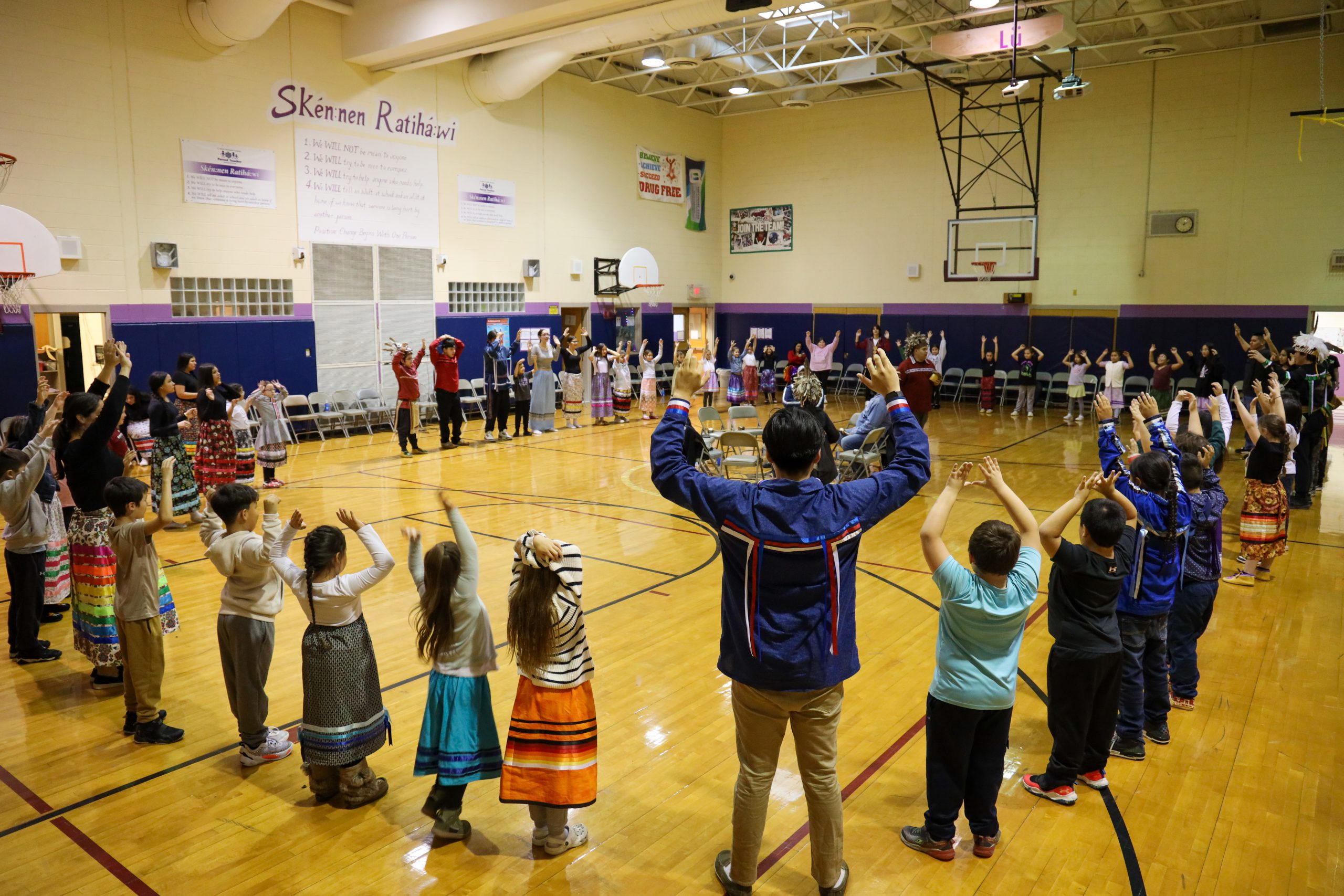 Dozens of students and staff standing in a circle in a gym holding up hands